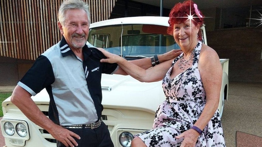 A man and woman dressed in 50s clothes stand in front of a vintage car, smiling and holding onto each other's shoulders