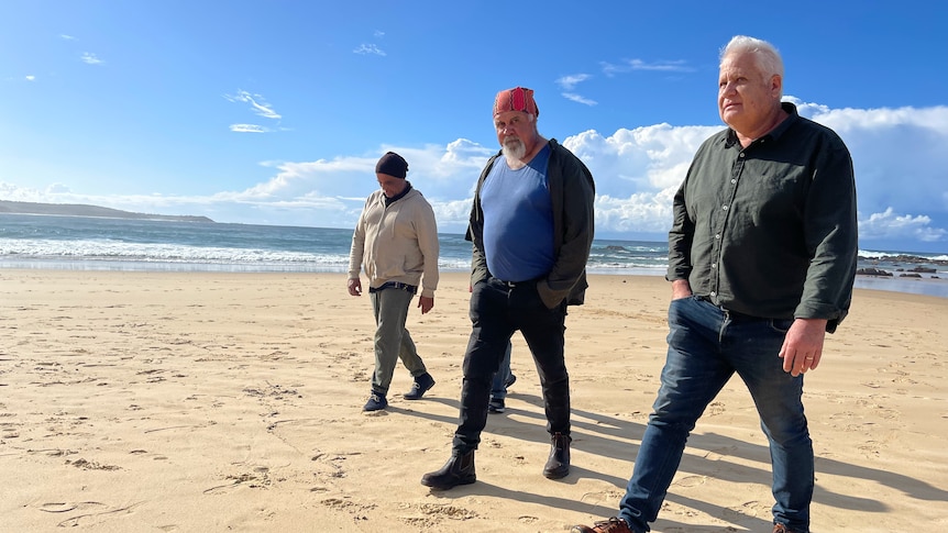 Three Yuin men walking along the beach wearing warm clothing.