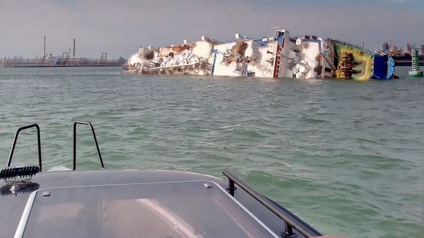 A ship half submerged in water with industrial buildings in the background