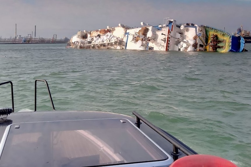 A ship half submerged in water with industrial buildings in the background