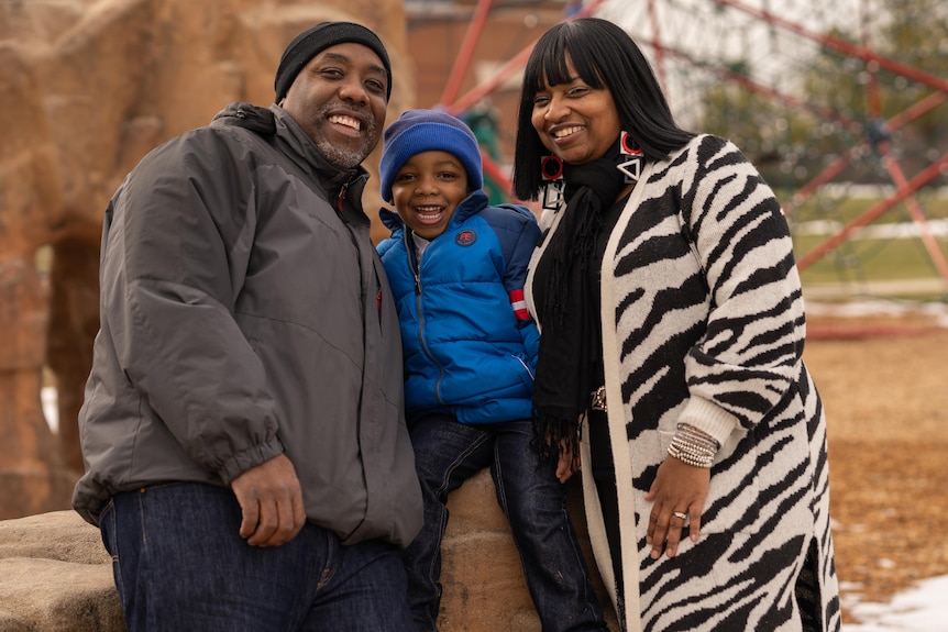 A man in a beanie and parka grins next to a small boy in a blue jacket, and a woman in a zebra print cardigan 