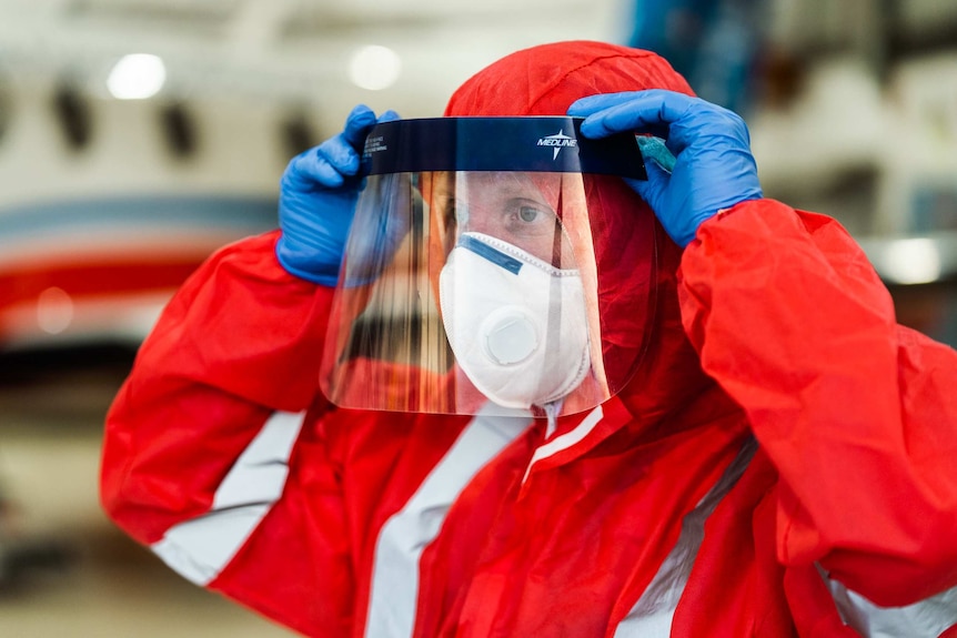 A close-up shot of a male doctor decked out in orange PPE putting a face visor on.