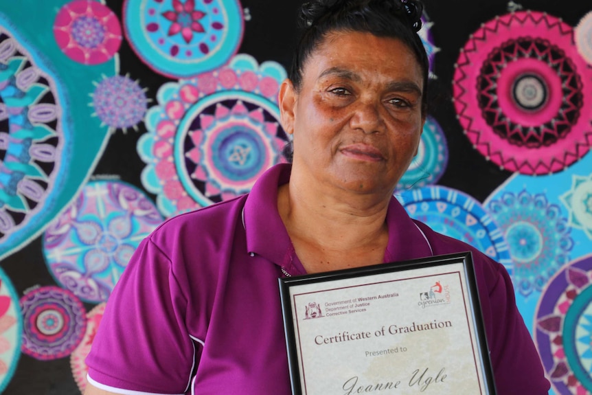 Joanne stands in front of a colourful mural while holding her graduation certificate.