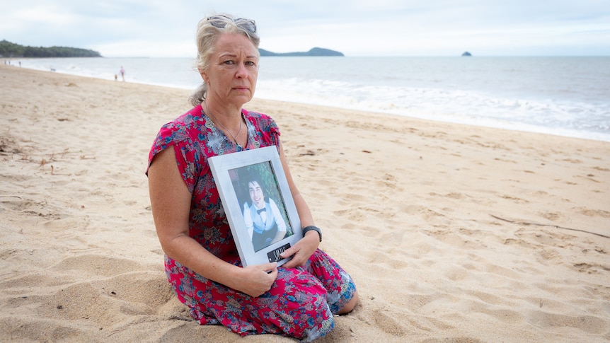 A middle aged woman sits on a beach holding a photo of her son. Her face is sad