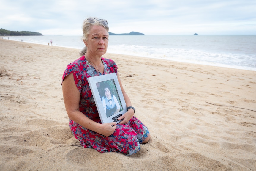 A middle aged woman sits on a beach holding a photo of her son. Her face is sad