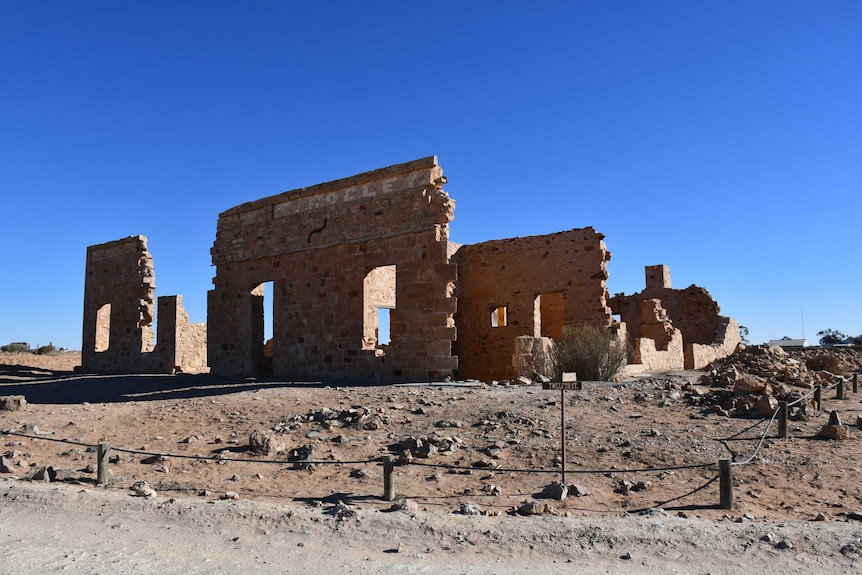 The stone walls of an old building cast a shadow onto the dirt it stands upon.