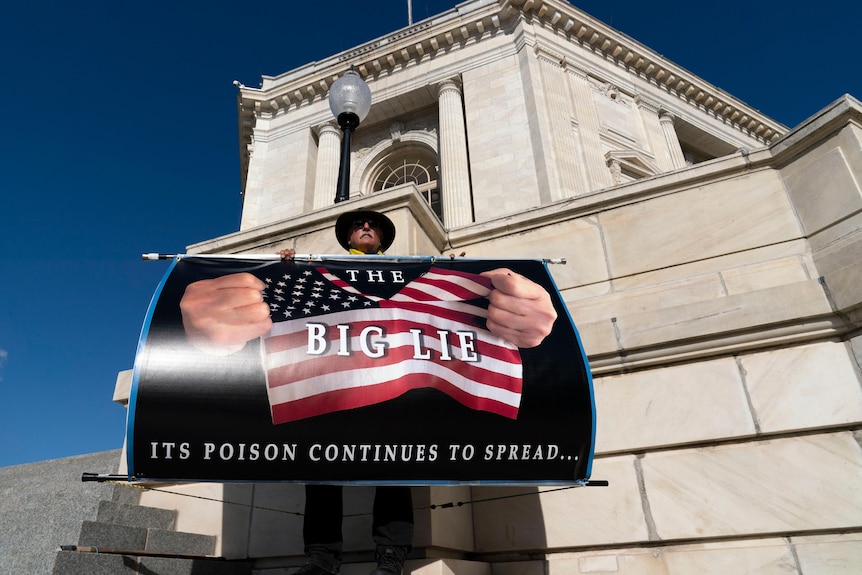 A man stands in front of a white building holding a sign, which says "The Big Lie: Its poison continues to spread..." 