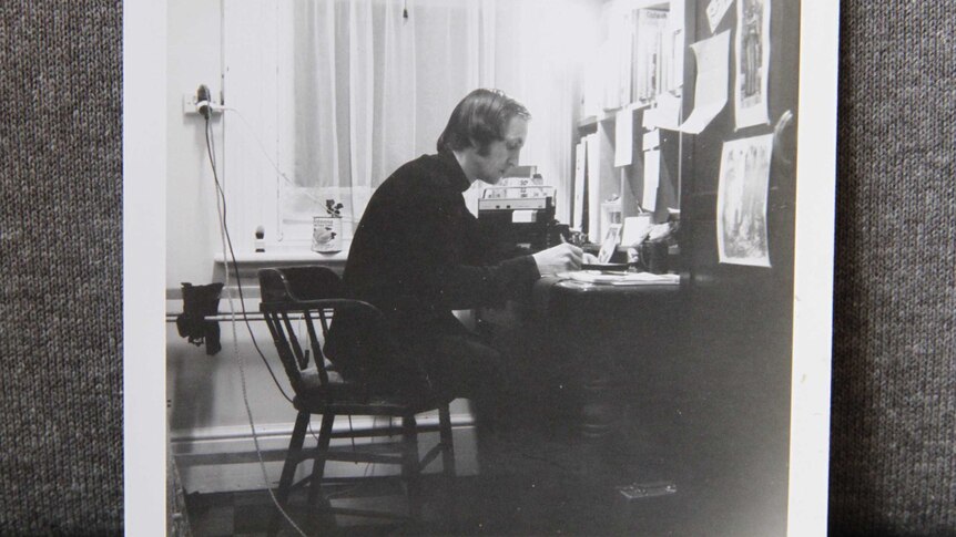 A black-and-white photo of a man sitting at a desk, writing on paper.