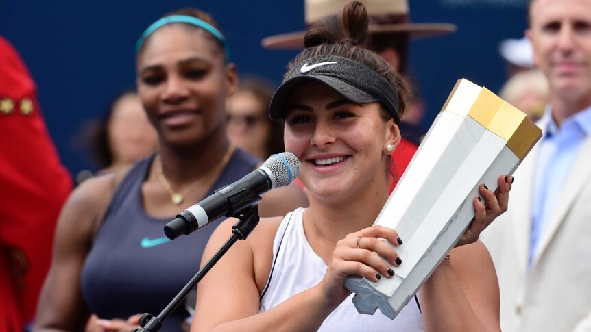 Bianca Andreescu holds up the white and gold Rogers Cup trophy. Serena Williams watches her from behind.