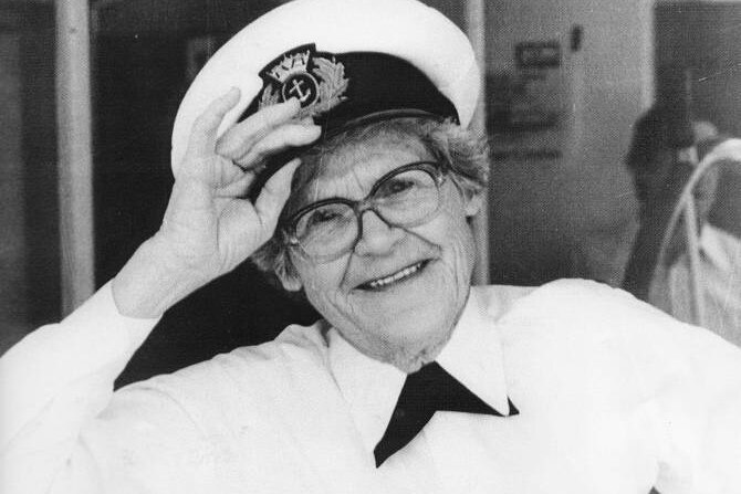 An older woman is holding her captain's hat and smiling. She is wearing a white shirt and dark pants.