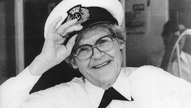 An older woman is holding her captain's hat and smiling. She is wearing a white shirt and dark pants.