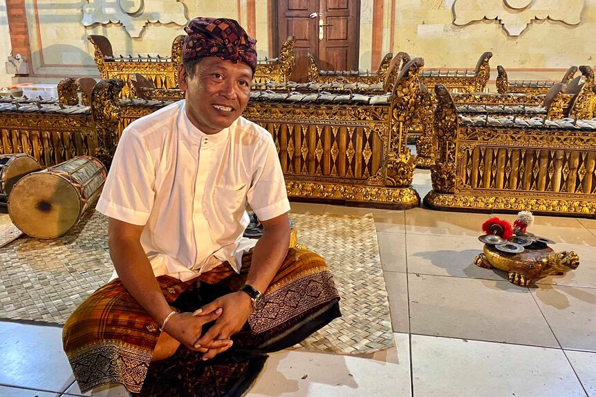 A man in traditional Balanese dress sits cross legged on a tiled floor with gilded musical instruments behind him