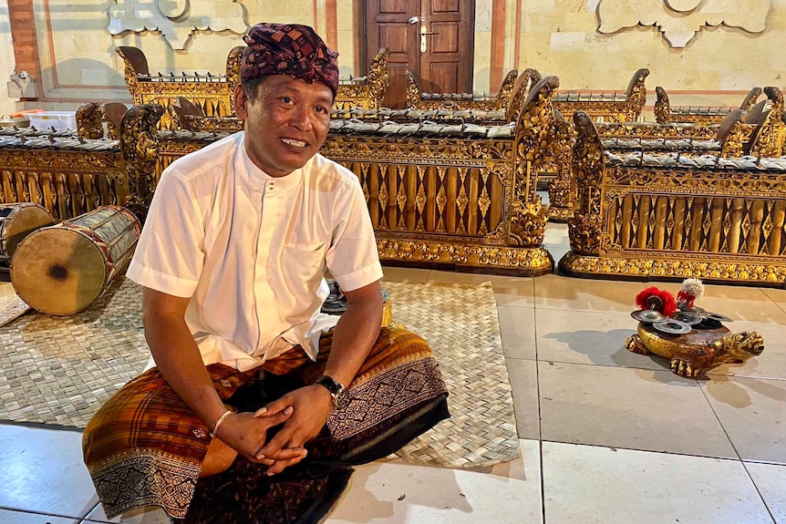 A man in traditional Balanese dress sits cross legged on a tiled floor with gilded musical instruments behind him