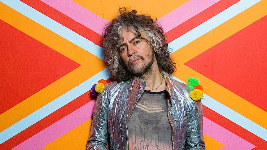 Wayne Coyne of The Flaming Lips wearing a silver, reflective jacket adorned with pom poms in front on a colourful background