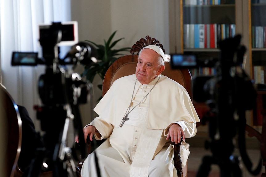 Pope Francis, sitting in a chair behind a video camera wearing papal garb, is illuminated by broadcast lights