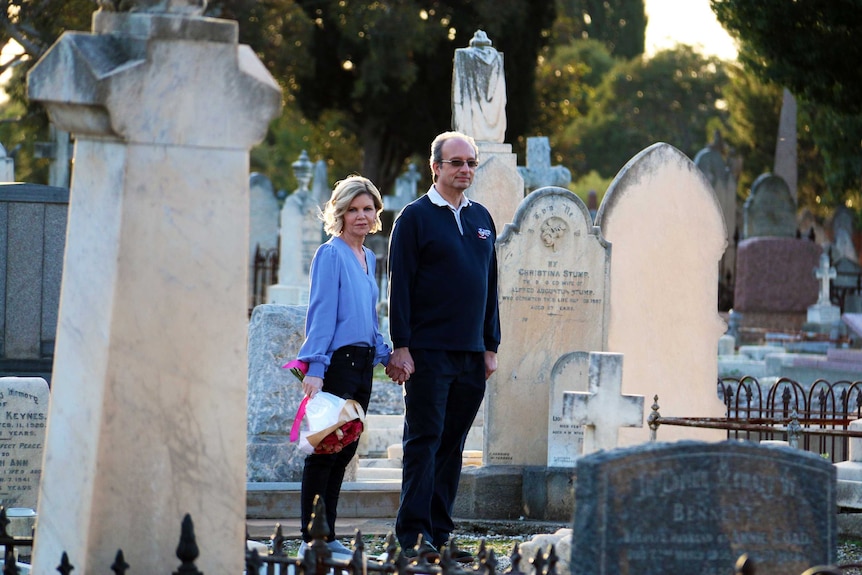 a man and a woman stand holding hands in a graveyard.  The woman is holding flowers