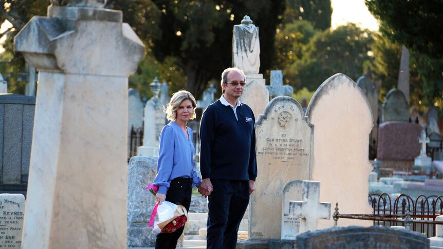 a man and a woman stand holding hands in a graveyard. The woman is holding flowers