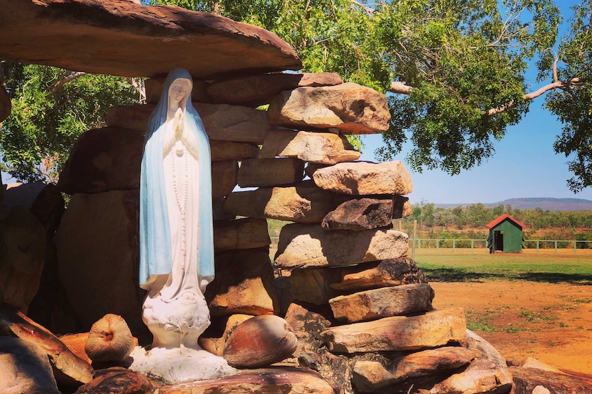 A small statue of Mary sits in a stone grotto with trees and a sunny sky in the background.