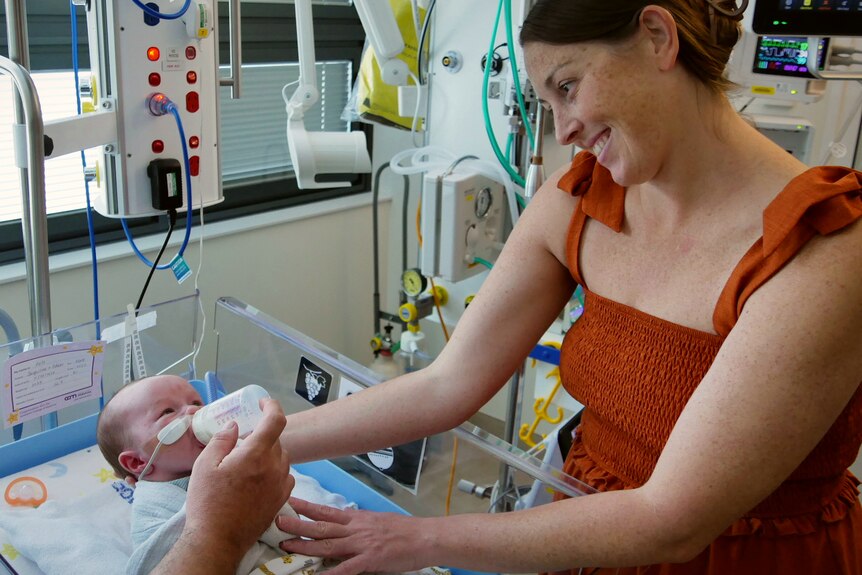 A man and a woman stand next to a baby in a cot in a hospital room