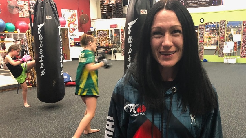 Sarah Lews with long black hair and hoodie top stands in front of two girls kicking punching bags