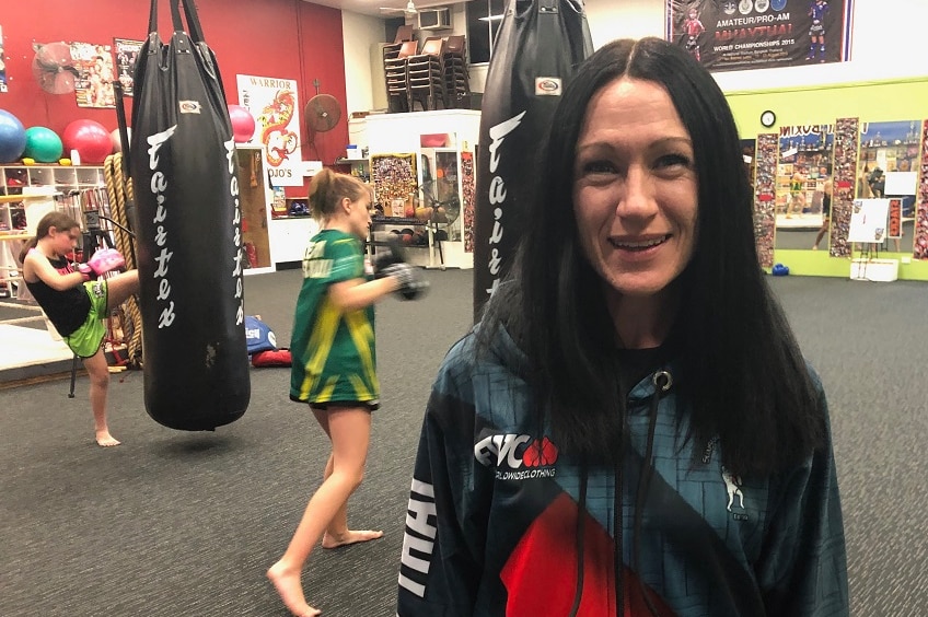 Sarah Lews with long black hair and hoodie top stands in front of two girls kicking punching bags