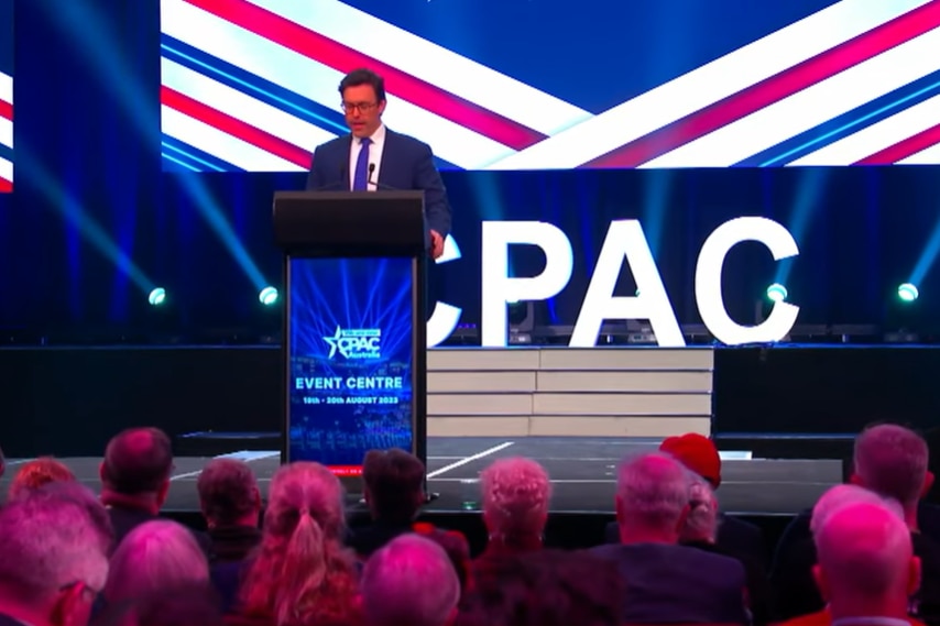 A man in a suit stands at a podium giving a speech in front of the CPAC logo on a stage