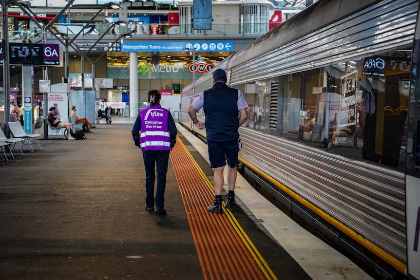 Two workers - one wearing a vline jacket - walking on a train platform beside a train