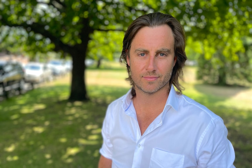 A man wearing a white shirt smiles at the camera while standing in a park.