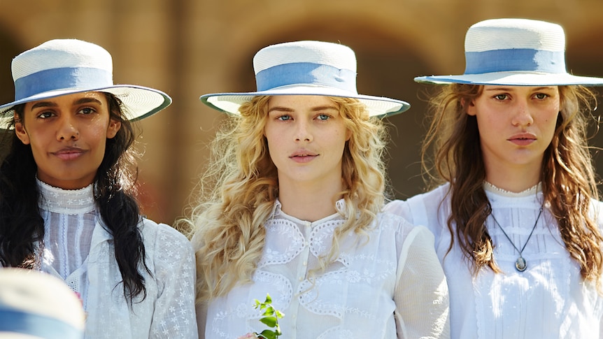 Three young women with long hair, wearing white straw hats with blue ribbons and white dresses in Victorian style.