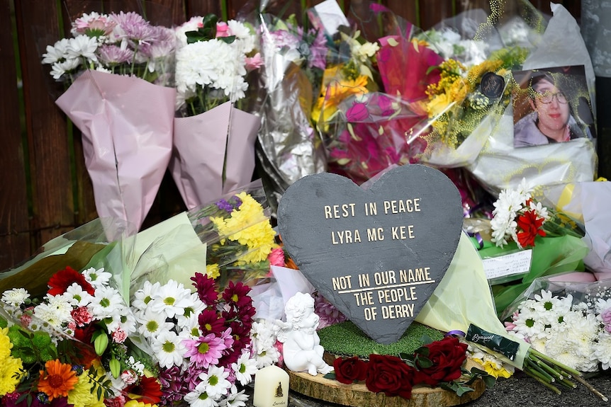 Bunches of flowers sit behind a heart-shaped memorial stone at a vigil on the side of a road