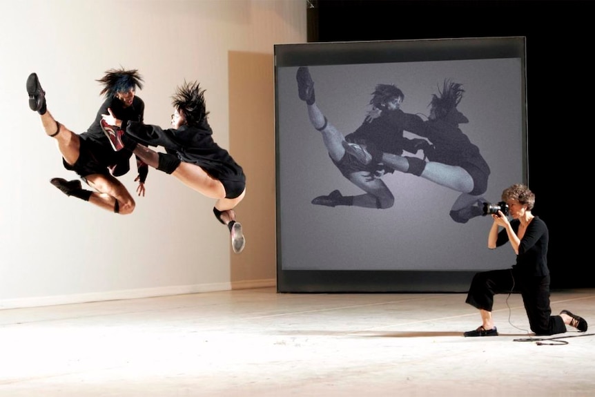 A photographer documents two dancers in the air on stage