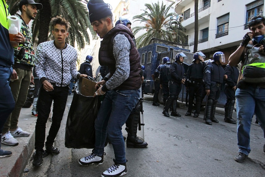 Two young men load rubbish into large garbage bags while police and others look on