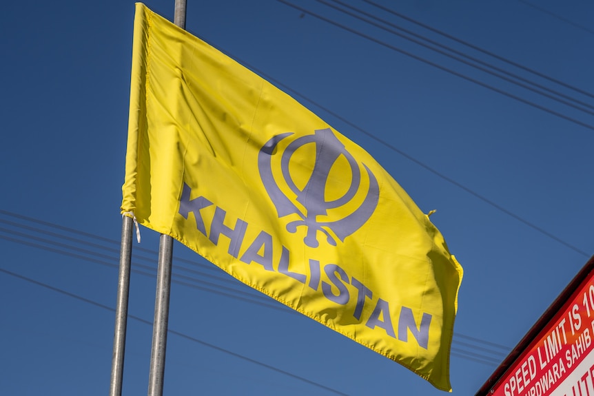 A yellow flag with a Khalistan logo and the word 'Khalistan' flaps against a blue sky.