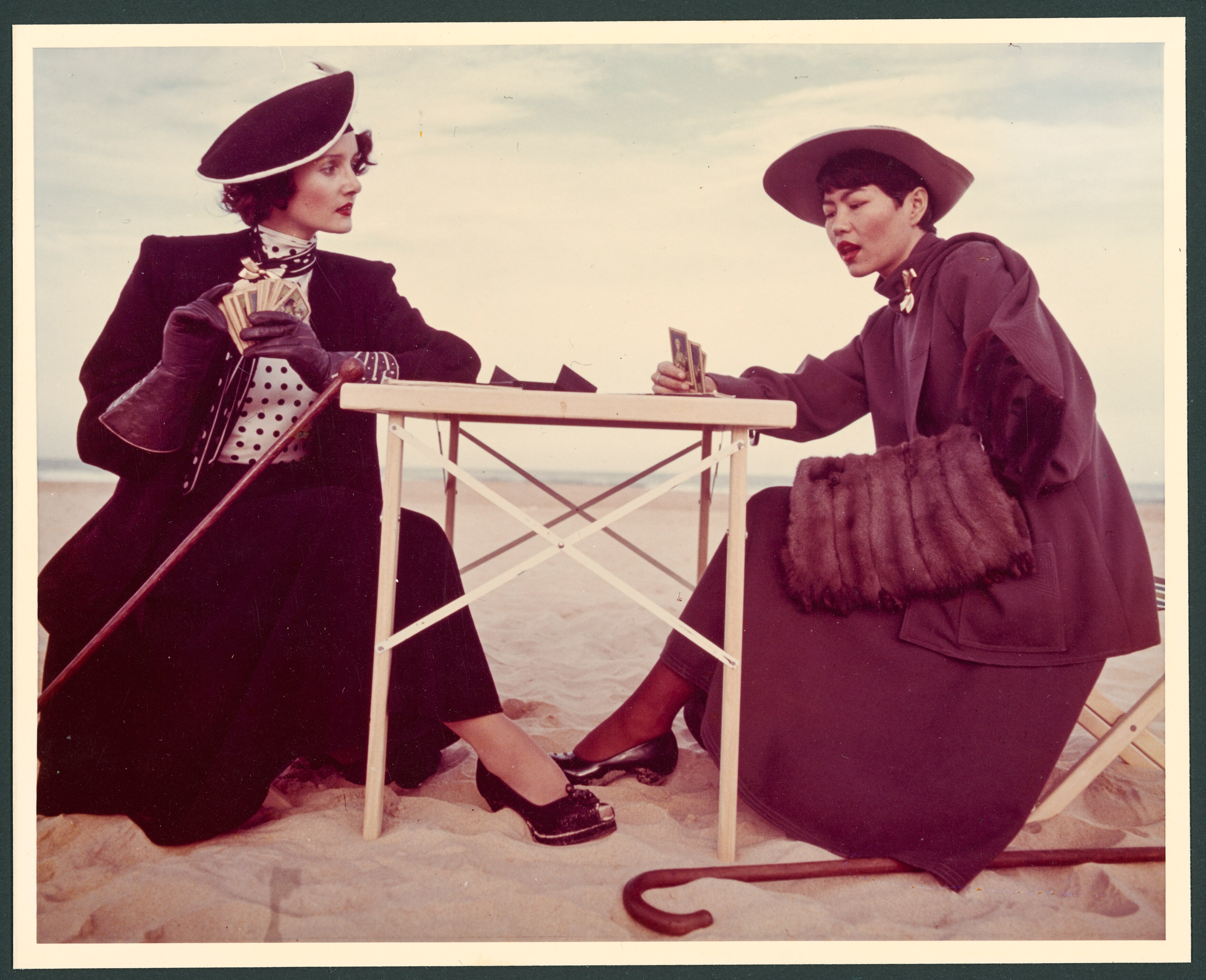 Two women wearing vintage clothes playing cards on a beach.