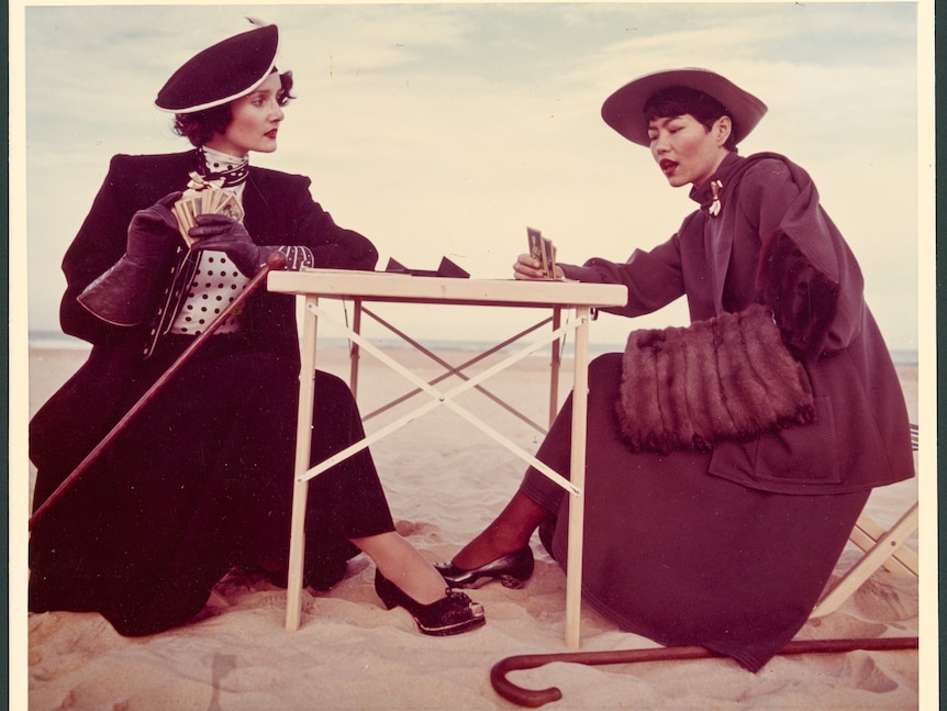 Two women wearing vintage clothes playing cards on a beach.