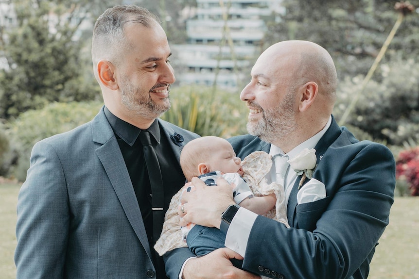 two men dressed in suits hold a newborn baby at a wedding