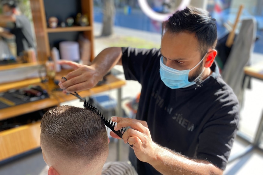 A man wearing a mask cuts a customer's hair.