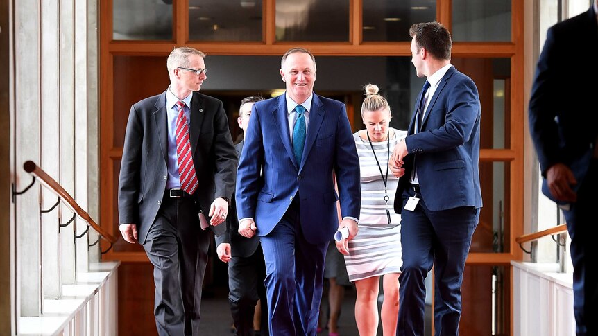 John Key smiles as walks down a corridor, flanked by a group of staffers.
