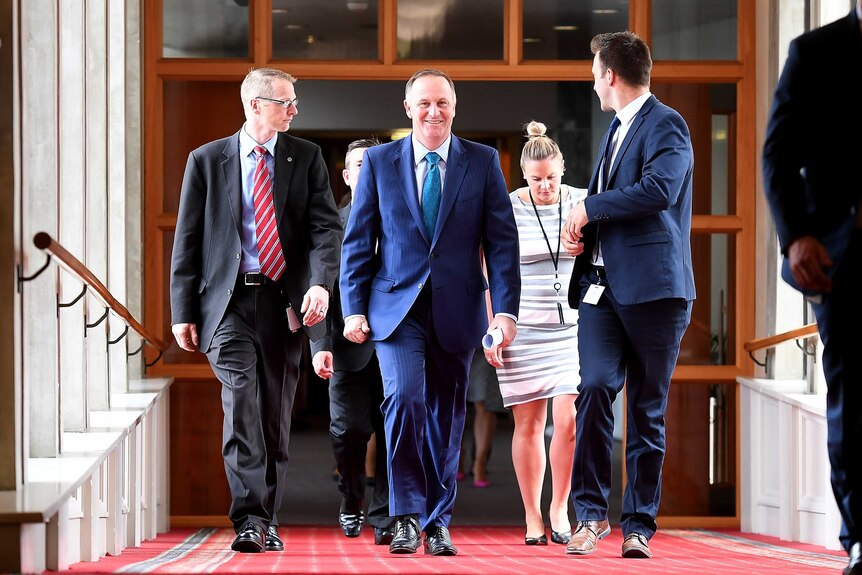 John Key smiles as walks down a corridor, flanked by a group of staffers.