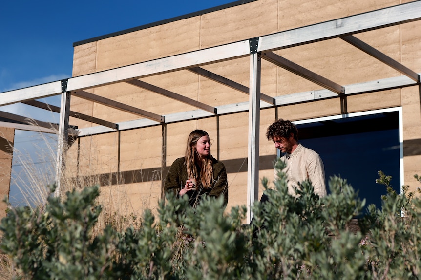 Woman wearing brown jumper sits on deck next to man wearing light coloured shirt in front of sandstone restaurant building