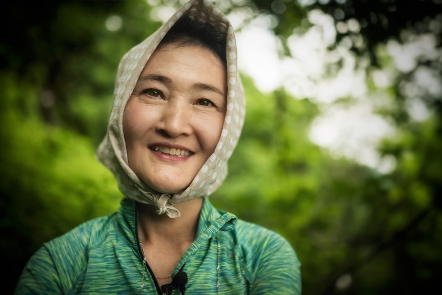A woman wearing a scarf tied around her head and a green shirt smiles at the camera.