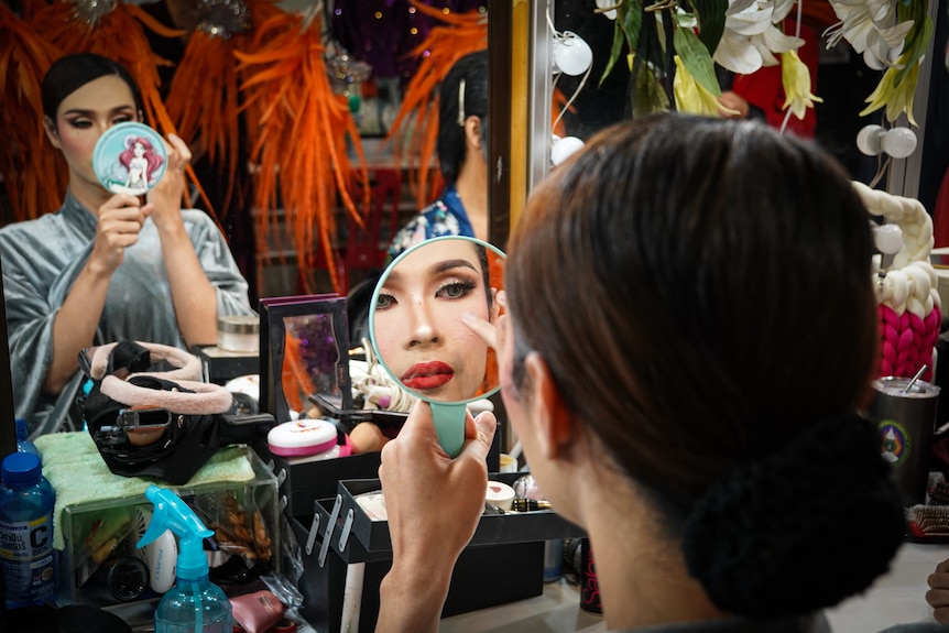 A woman holding up a small mirror to apply make up in front of a bigger mirror