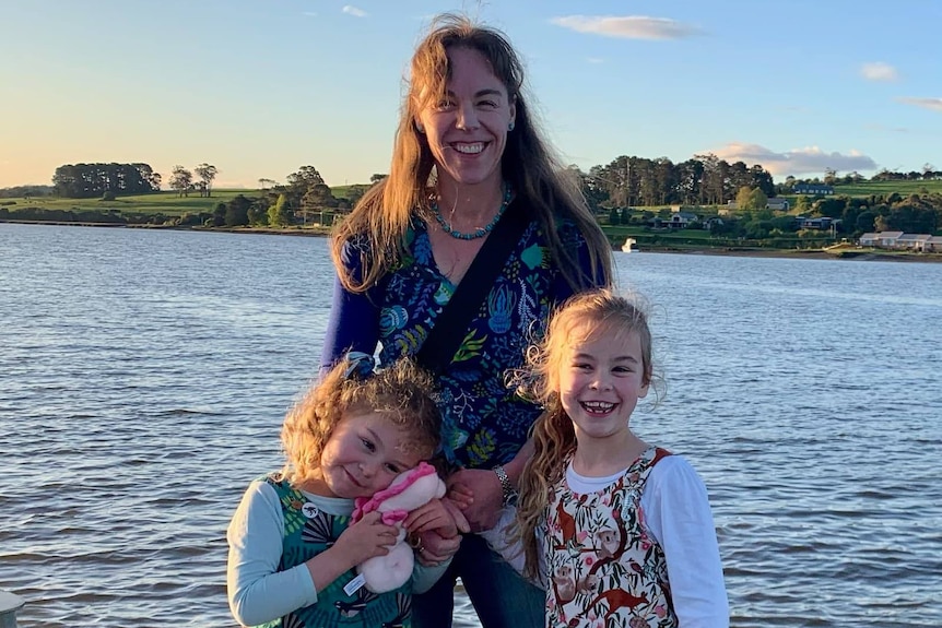 A woman smiling with two small children. They're all holding hands.