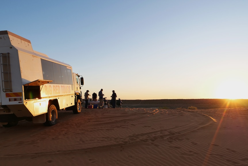 A tour bus parked on a red sand dune with people standing around and the sun setting in front. 