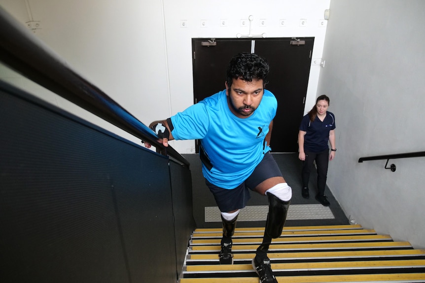 A man with prosthetic legs climbs an indoor staircase while holding the handrail