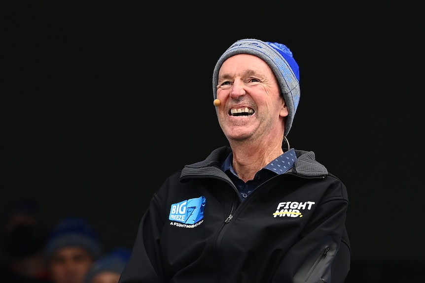 A man wearing a blue-and-grey beanie and a headset mike grins at the crowd during an MND fundraising event at the MCG
