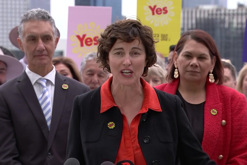 A woman wearing a black jacket over a red blouse speaks at a press conference. 