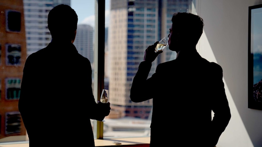 Two men drink champagne while standing at an office window.