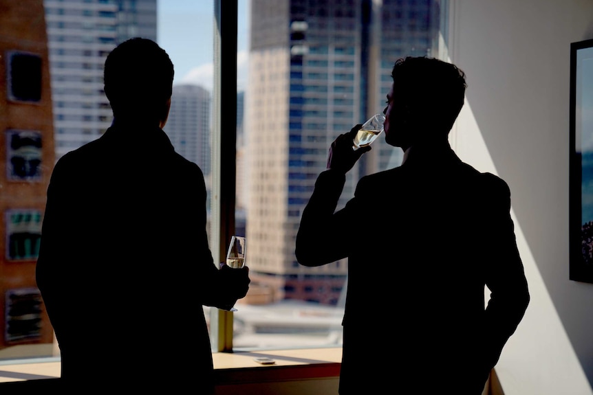 Two men drink champagne while standing at an office window.