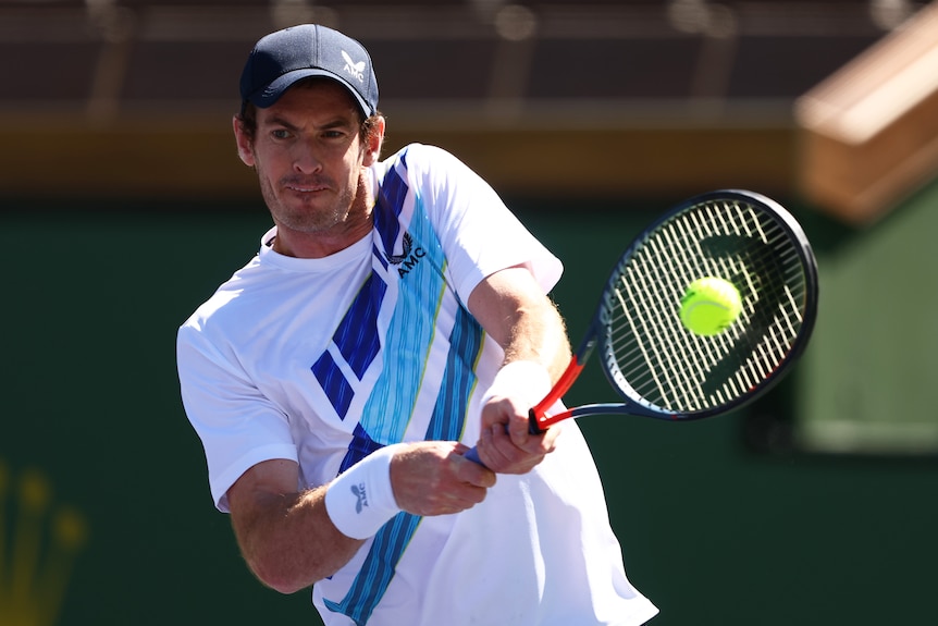 A tennis player grimaces as his racquet connects with the ball for a two-handed backhand during a match. 
